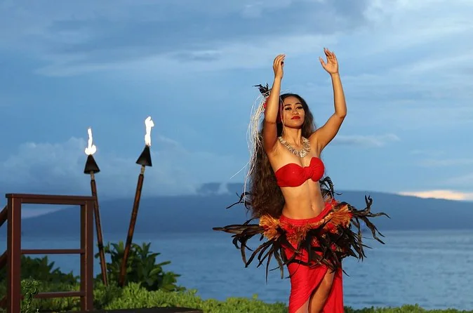 A dancer in a red costume between tiki torches on an oceanfront lawn