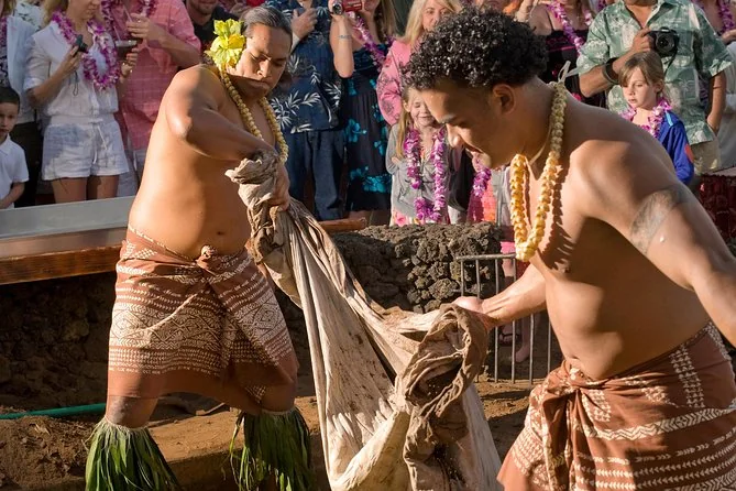 Two performers lifting cloth off the imu earth oven as guests wearing lei watch