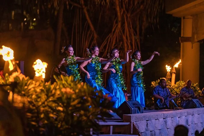 Dancers in blue dresses and green lei on a torch-lit stage with musicians
